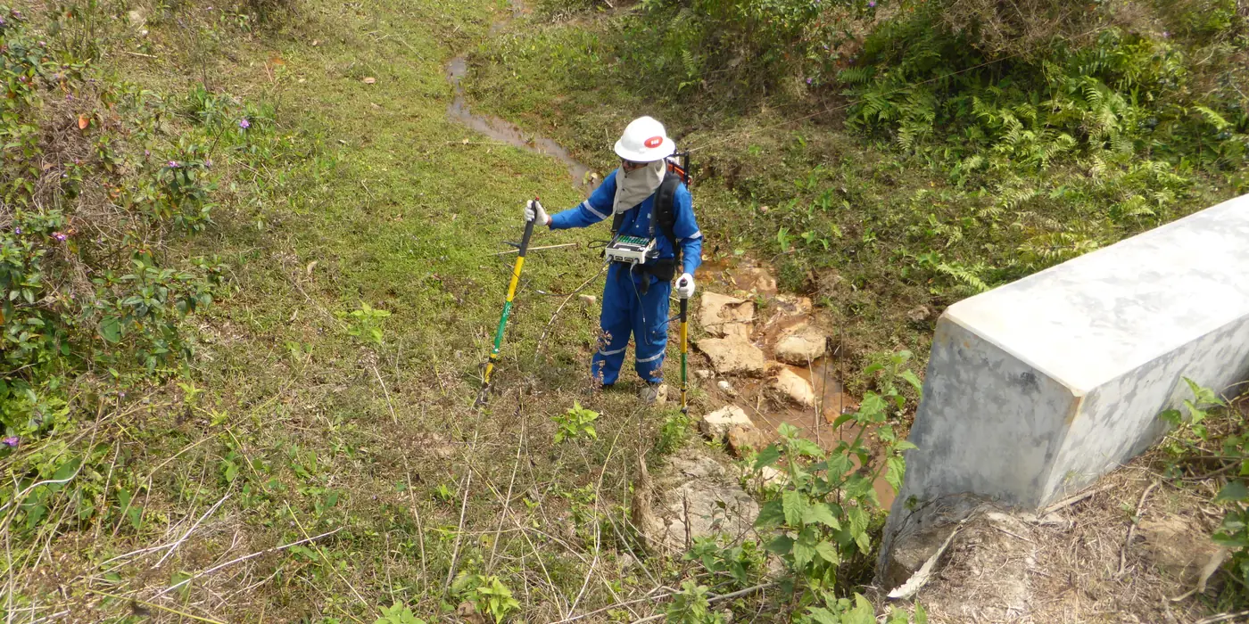 JST surveyor walking a pipeline right-of-way with DCVG probes, between a concrete marker and the next test post